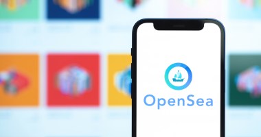 Celular à frente de grande tela de computador mostra logo da Opensea