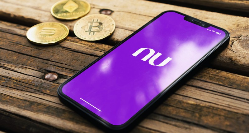 Sob um banco de madeira três criptomoedas ao lado de um celular com logo do Nubank