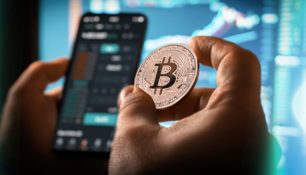 mãos segurando celular com grafico e moeda de Bitcoin