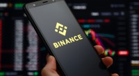 Mão segurando um celular com o logo da Binance em frente a uma tela com um livro de ofertas de criptomoedas