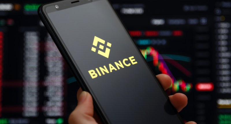 Mão segurando um celular com o logo da Binance em frente a uma tela com um livro de ofertas de criptomoedas