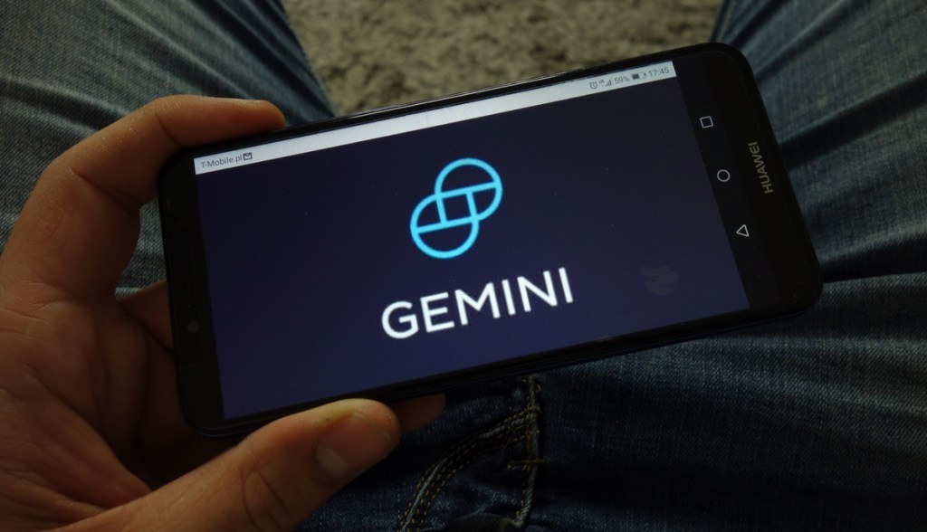 Mão segurando um celular com o logo da corretora Gemini