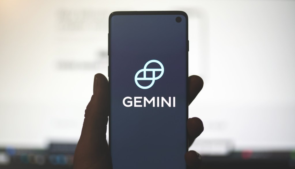 Mão segurando um celular com o logo dourado da corretora Gemini