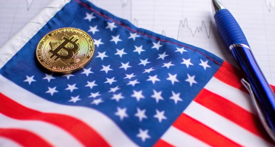 Moeda dourada de Bitcoin sobre bandeira dos Estados Unidos ao lado de uma caneta esferográfica