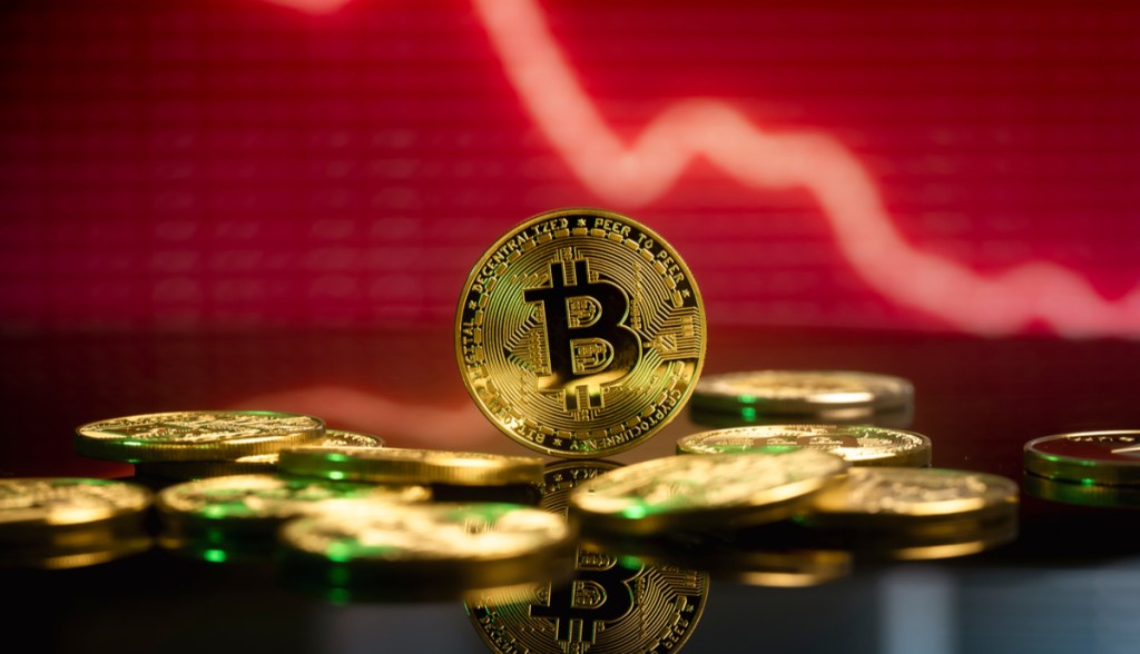 Moedas douradas de Bitcoin sobre uma mesa à frente de um gráfico de preço em vermelho, indicando uma drástica queda