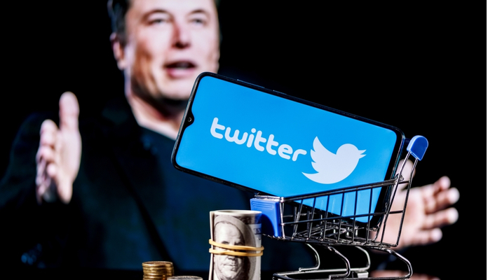Elon Musk é retratado ao fundo. No primeiro plano, um carrinho de compras contendo o logo do Twitter.