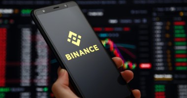 mão segurando celular com logo da binance e gráficos ao fundo