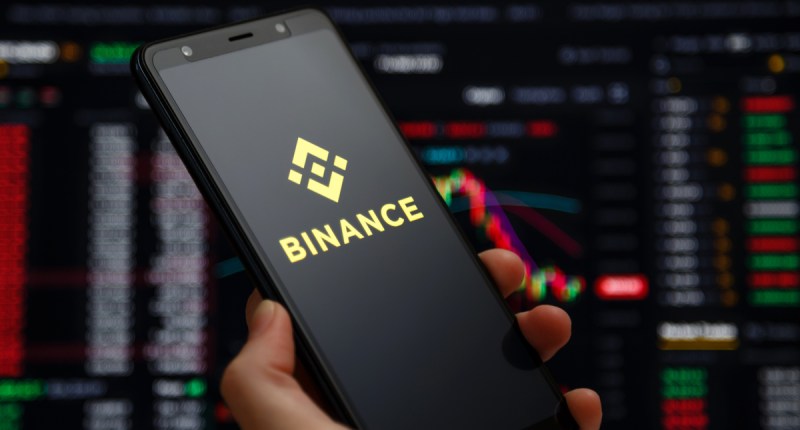 mão segurando celular com logo da binance e gráficos ao fundo