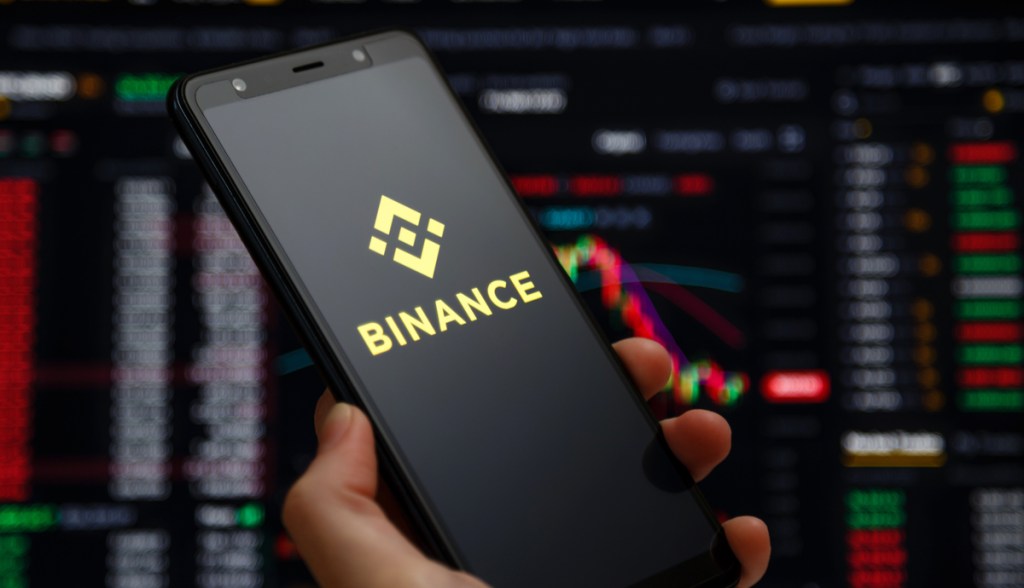 mão segurando celular com logo da binance e gráficos ao fundo