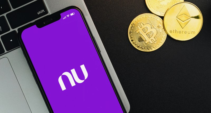 Foto com um celular aberto no aplicativo Nubank, e criptomoedas ao redor.