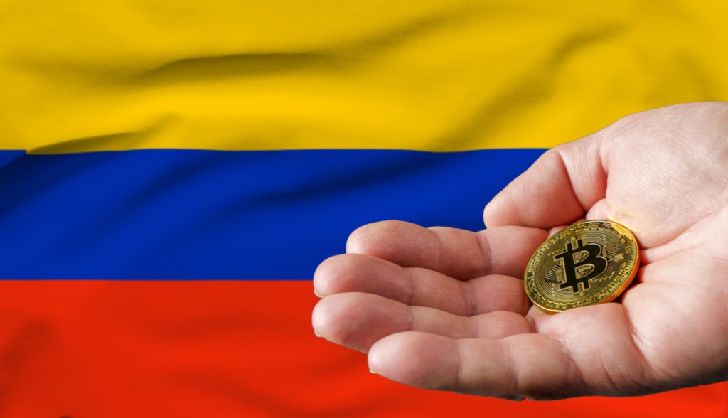 Mão segurando uma moeda dourada de Bitcoin (BTC) em frente à bandeira da Colômbia