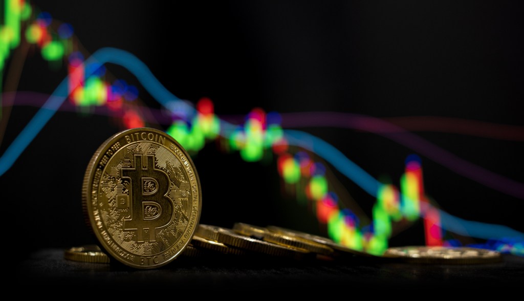 Moeda dourada de Bitcoin com gráfico de candles indicando drástica queda ao fundo