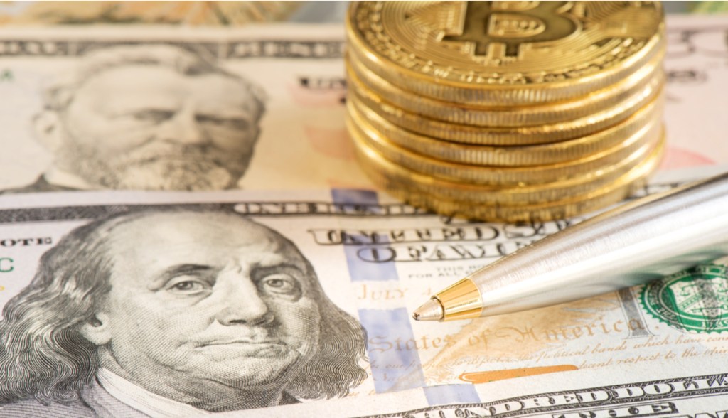 Pilhas de moedas douradas de Bitcoin (BTC) sobre cédulas de dólares e próximas a uma caneta esferográfica