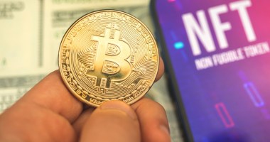 Dedos seguram moeda de bitcoin ao lado de tela que mostra sigla NFT