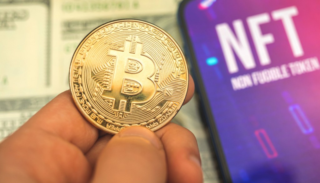 Dedos seguram moeda de bitcoin ao lado de tela que mostra sigla NFT