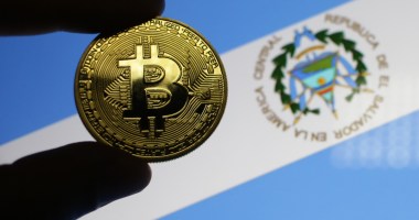 Pessoa segura moeda de bitcoin à frente de bandeira de El Salvador