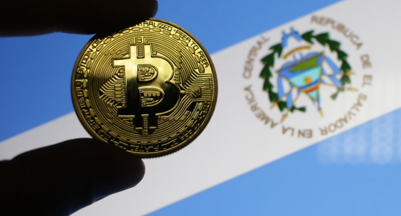 Pessoa segura moeda de bitcoin à frente de bandeira de El Salvador