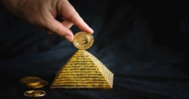 uma pessoa apoia uma moeda de bitcoin no topo de uma maquete de pirâmide dourada