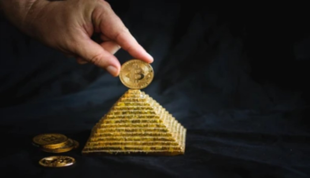 uma pessoa apoia uma moeda de bitcoin no topo de uma maquete de pirâmide dourada