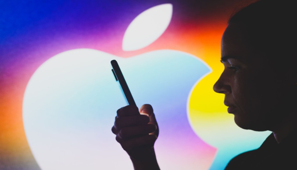Pessoa segura um smartphone à frente de um logo da Apple grande e colorido