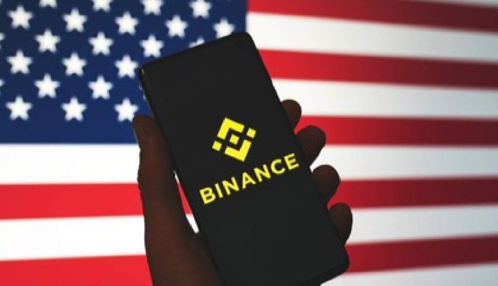 celular com logo da binance e bandeira dos EUA ao fundo