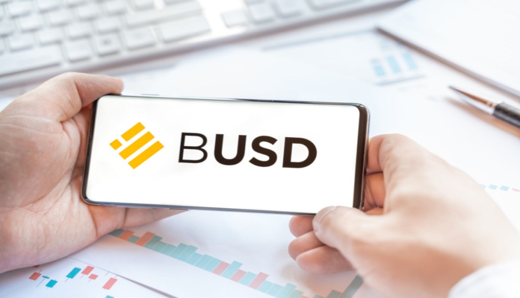 Pessoa segura smartphone com logo BUSD com as mãos apoiado em uma mesa