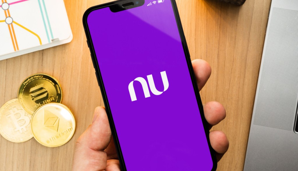 Celular mostra logotipo Nubank - abaixo mesa com moedas de bitcoin, ethereum e solana
