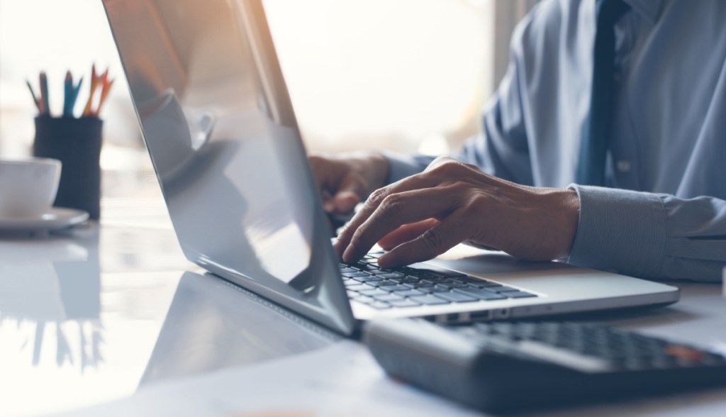 Pessoa digita em um laptop - ao lado uma calculadora