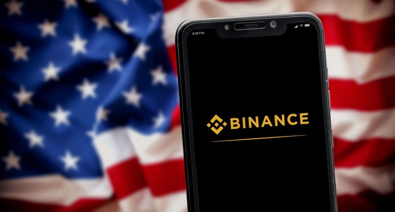 logo a binance no celular e bandeira dos EUA no fundo