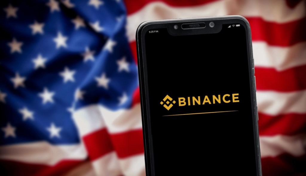 logo a binance no celular e bandeira dos EUA no fundo