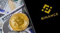 Celular com logo da Binance, moedas de Bitcoin e ETeherem em cima de notás de cem dólares