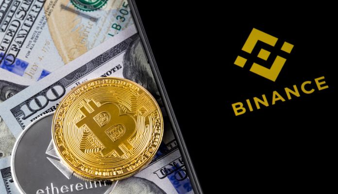 Celular com logo da Binance, moedas de Bitcoin e ETeherem em cima de notás de cem dólares