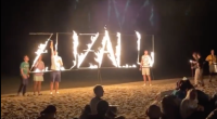 Letreiro "Zuzalu" em chamas em uma praia com diversos participantes