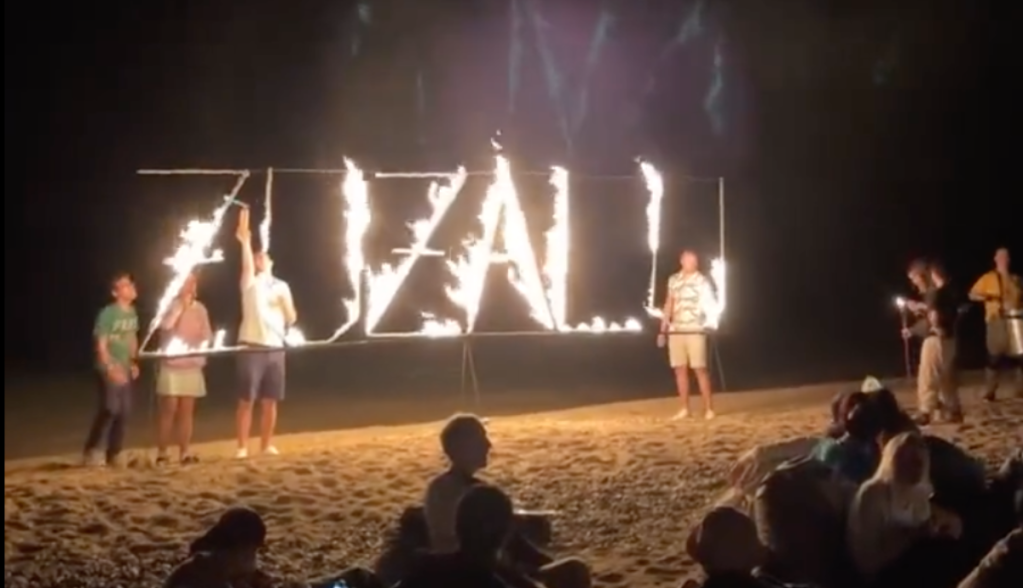 Letreiro "Zuzalu" em chamas em uma praia com diversos participantes