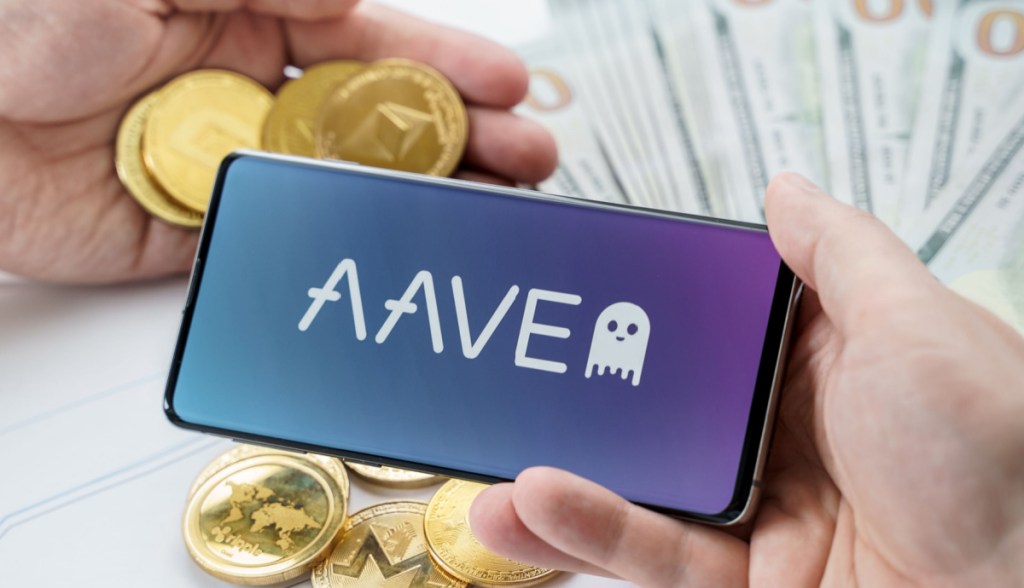 Uma mão segunra smartphone comlogomarca Aave e outra mãos critpomeodas Ethereum