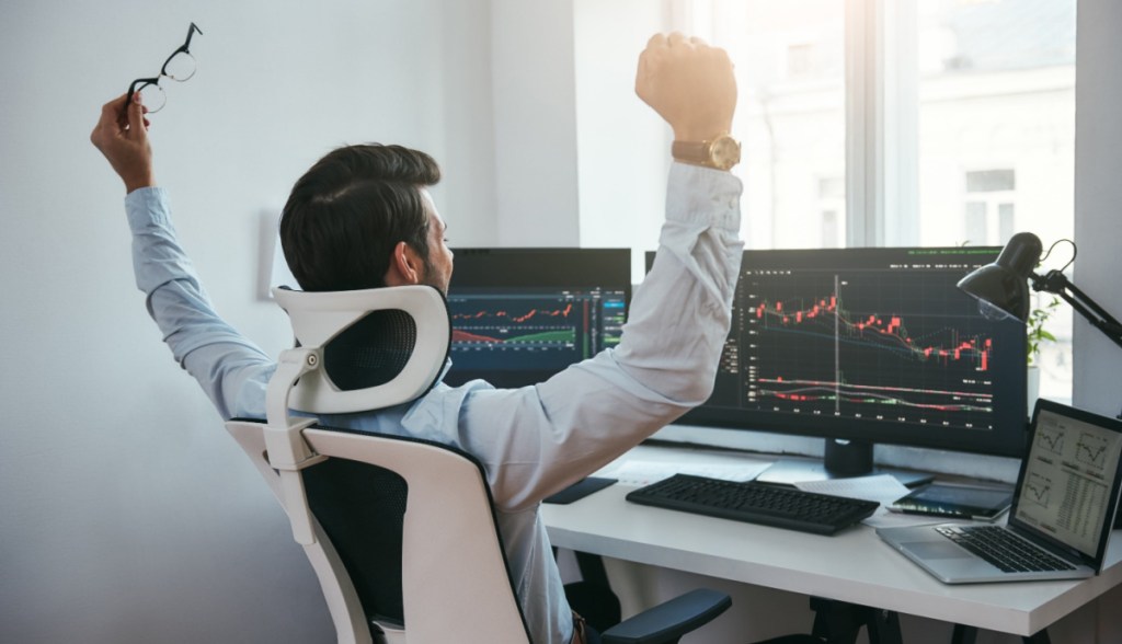trader com braços levantados em frente a computador com graficos