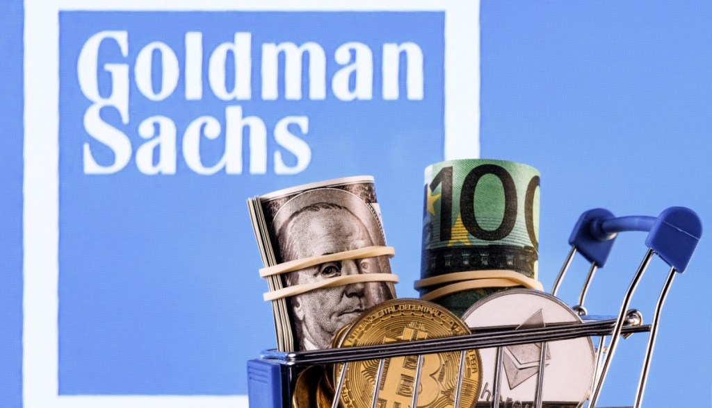 carrinho de compras com criptomoedas e dinheiro; ao fundo, logo do Goldman Sachs