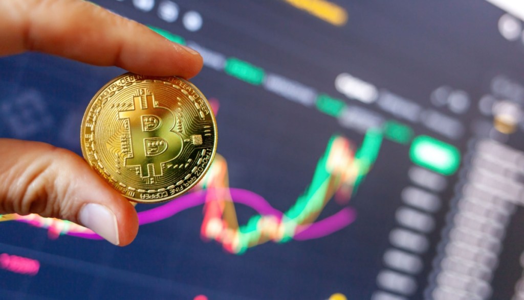 dedos seguram moeda de Bitcoin à frente de gráfico financeiro